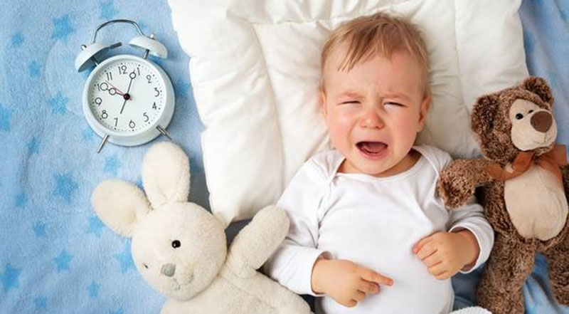 A baby lying on a bed cries while surrounded by a white bunny plush, a brown teddy bear, and a blue alarm clock on a light blue star-patterned blanket.