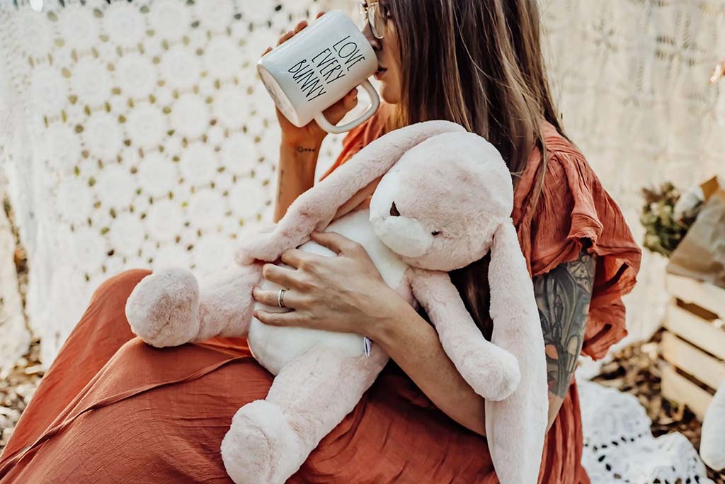 Woman relaxing indoors while holding a soft pink bunny plush and drinking from a mug, showing the comforting and emotional role of plush toys in everyday life.