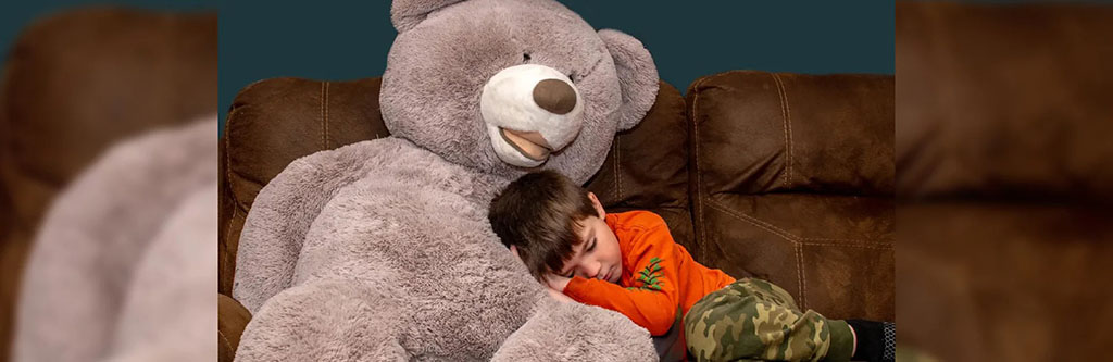A young child resting against an oversized plush teddy bear on a sofa, showing the scale, softness, and comfort appeal of large stuffed animals designed for emotional comfort, relaxation, and cozy home environments.