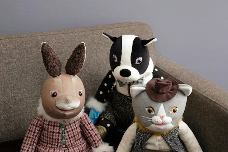 Three vintage-style plush animals sitting on a sofa, including a rabbit, a dog, and a cat dressed in classic clothing