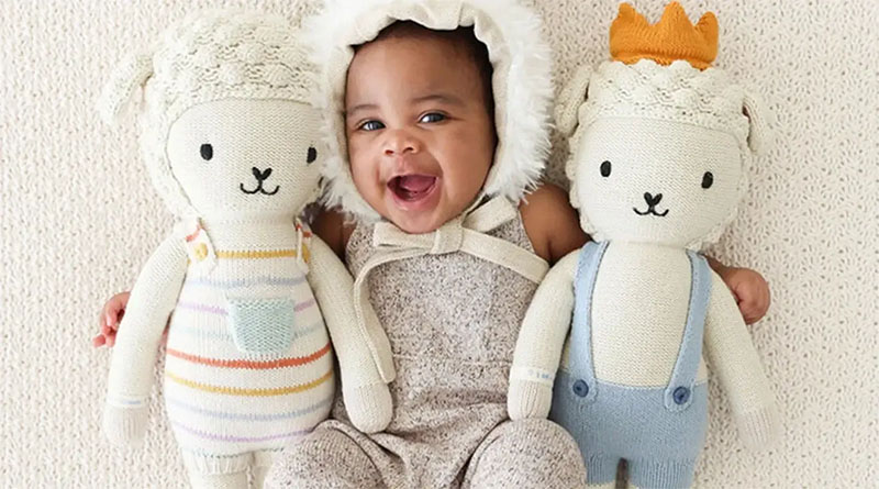 Smiling baby lying between two knitted plush dolls on a soft blanket