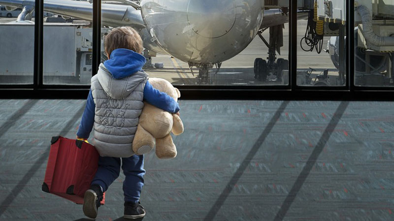 Young child at an airport holding a stuffed teddy bear while looking at an airplane through a large window