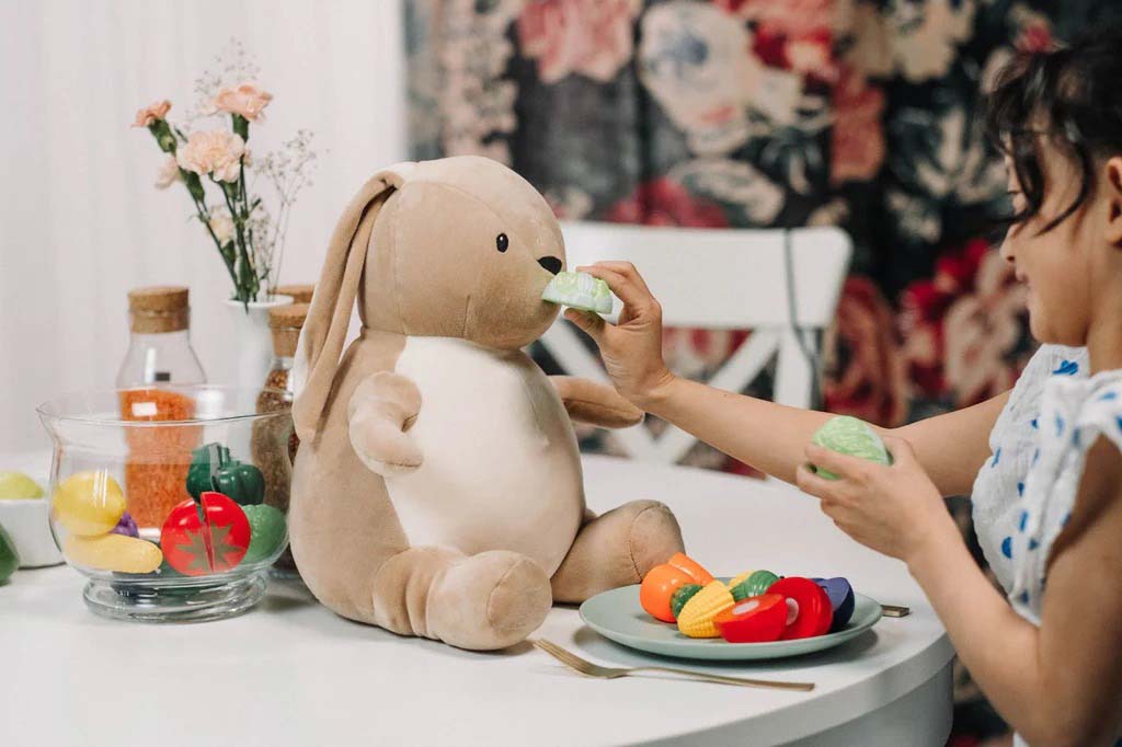 A young child pretending to feed a plush bunny toy with colorful toy vegetables at a small table during imaginative play