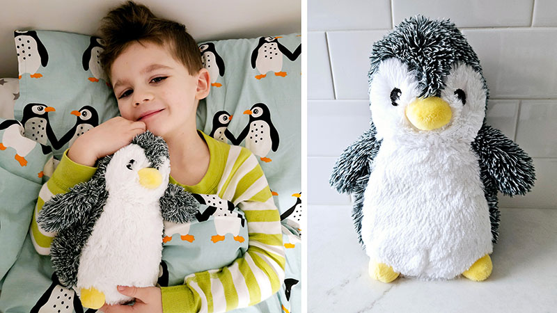 Child cuddling a weighted penguin stuffed animal in bed next to a standalone penguin plush on a white surface