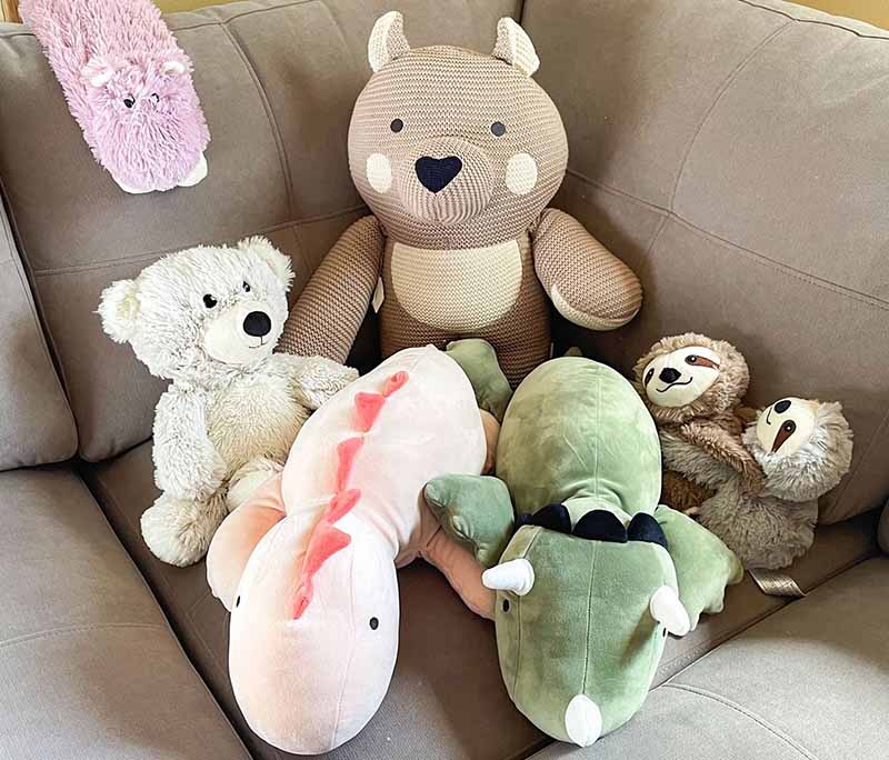 Assorted plush stuffed animals arranged together on a sofa, including a teddy bear, sloth, dinosaur, and soft animal pillows