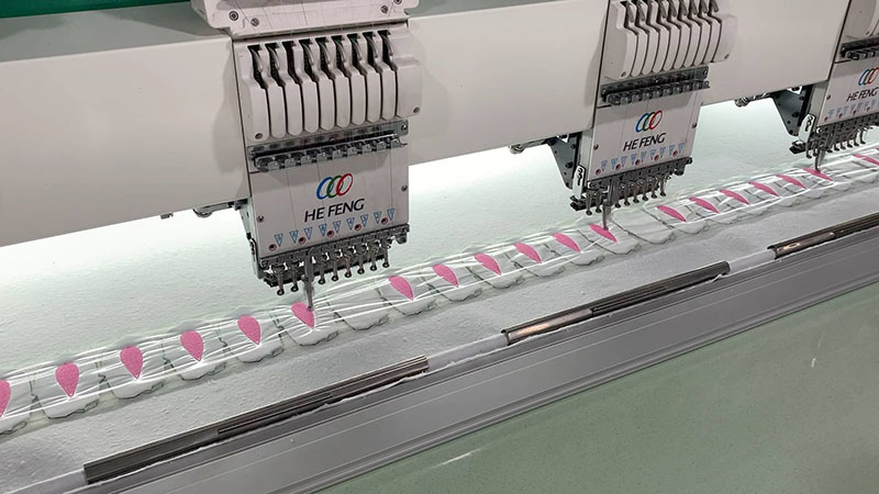 Industrial multi-head embroidery machines stitching repeating pink patterns onto plush fabric strips