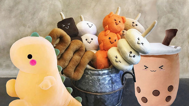 A collection of cute novelty plush toys arranged in a metal bucket, including kawaii-style food characters and a pastel dinosaur plush
