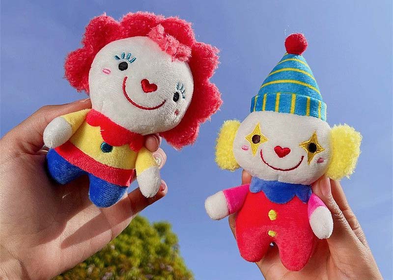 Two colorful clown plush toys held in hands outdoors, featuring smiling faces, bright outfits, and soft textured fabric