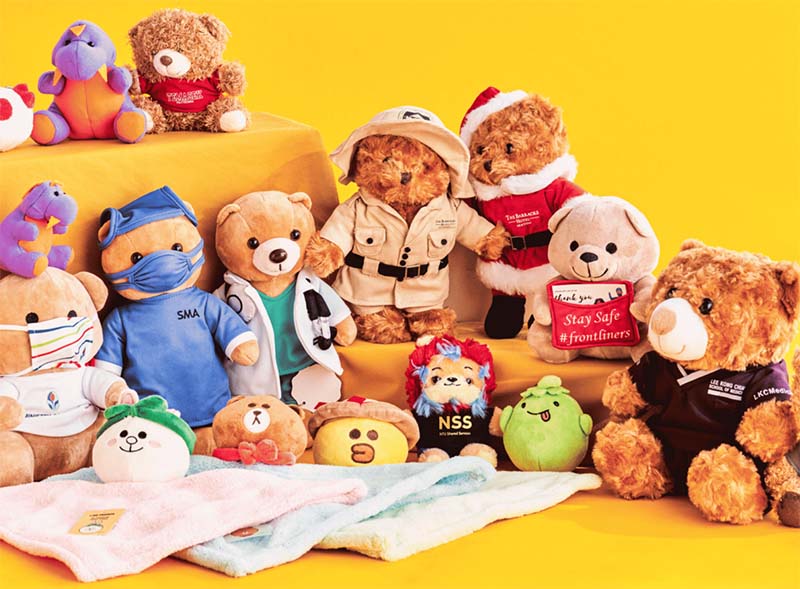 A collection of custom teddy bears dressed in themed outfits, including medical, explorer, holiday, and branded uniforms, arranged together on a bright yellow background