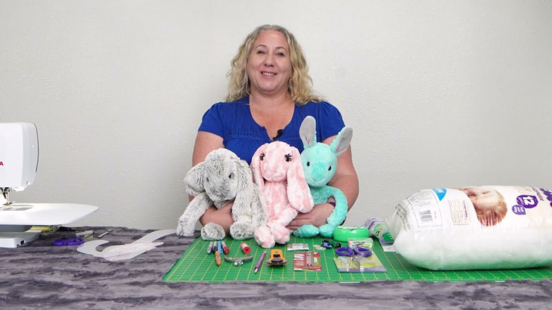 Woman sitting at a sewing table with handmade plush bunny toys, sewing tools, and stuffing materials laid out in front
