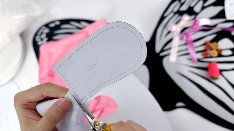 Hands cutting a paper plush toy pattern with scissors, placed over black and pink fabric pieces, with sewing tools and small accessories visible in the background during the plush-making process.