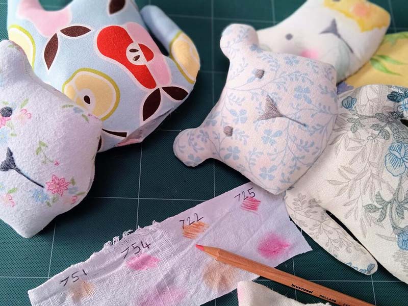 Close-up of handmade fabric plush dolls in soft pastel patterns lying on a cutting mat, with a fabric color swatch strip labeled with pencil markings and a colored pencil used for testing fabric dyes.