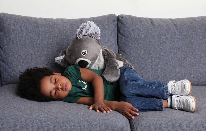 Toddler sleeping on a gray sofa with a weighted stuffed animal resting on their back