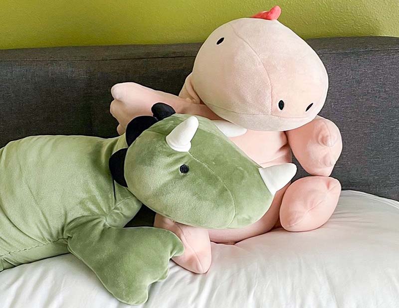 Two large plush dinosaurs resting on a bed, one green and one pink, designed as soft weighted stuffed animals for comfort and relaxation