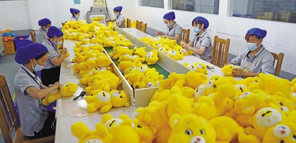Workers wearing masks assembling and checking yellow teddy bear plush toys on a factory production line.