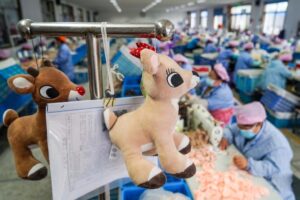 Deer plush toys hanging on a production rack with paperwork, with workers sewing in a busy plush toy factory background.