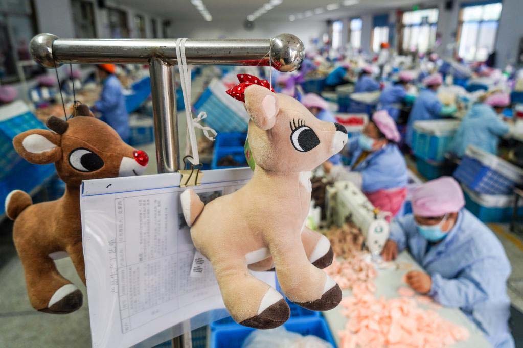 Deer plush toys hanging on a production rack with paperwork, with workers sewing in a busy plush toy factory background.