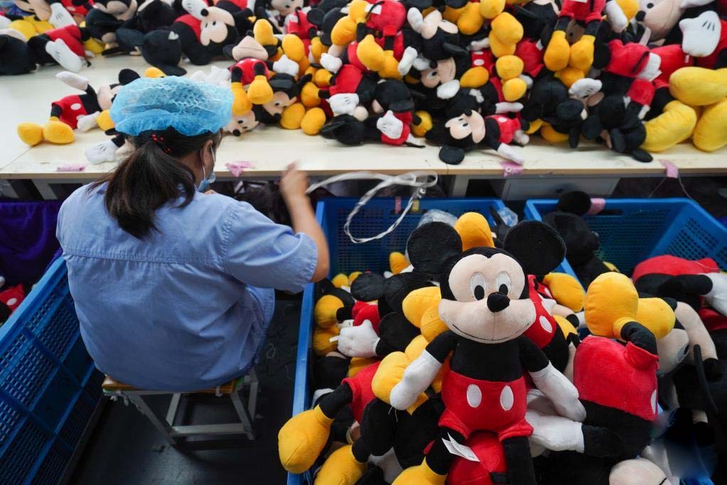 Factory worker sorting piles of cartoon character plush toys on a production line with bins full of stuffed dolls.