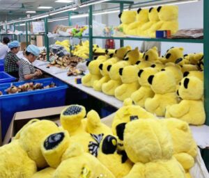 Workers assembling and sorting yellow plush toys on a factory production line with shelves full of stuffed animals.