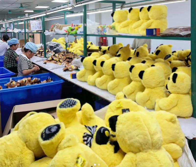 Workers assembling and sorting yellow plush toys on a factory production line with shelves full of stuffed animals.