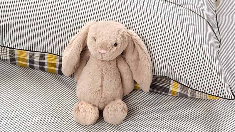 Beige plush bunny with floppy ears sitting on a bed against patterned pillows
