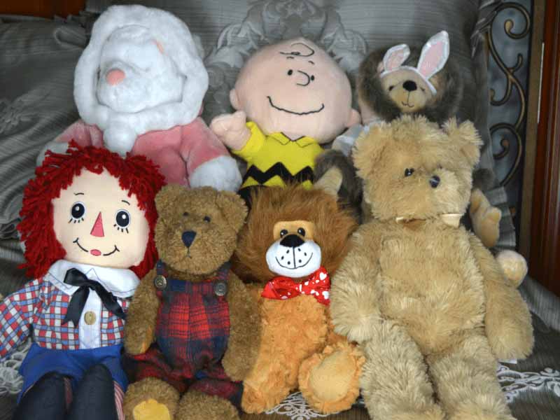 Group of plush toys on a couch, including teddy bears, a rag doll, a bunny, and a cartoon character doll.