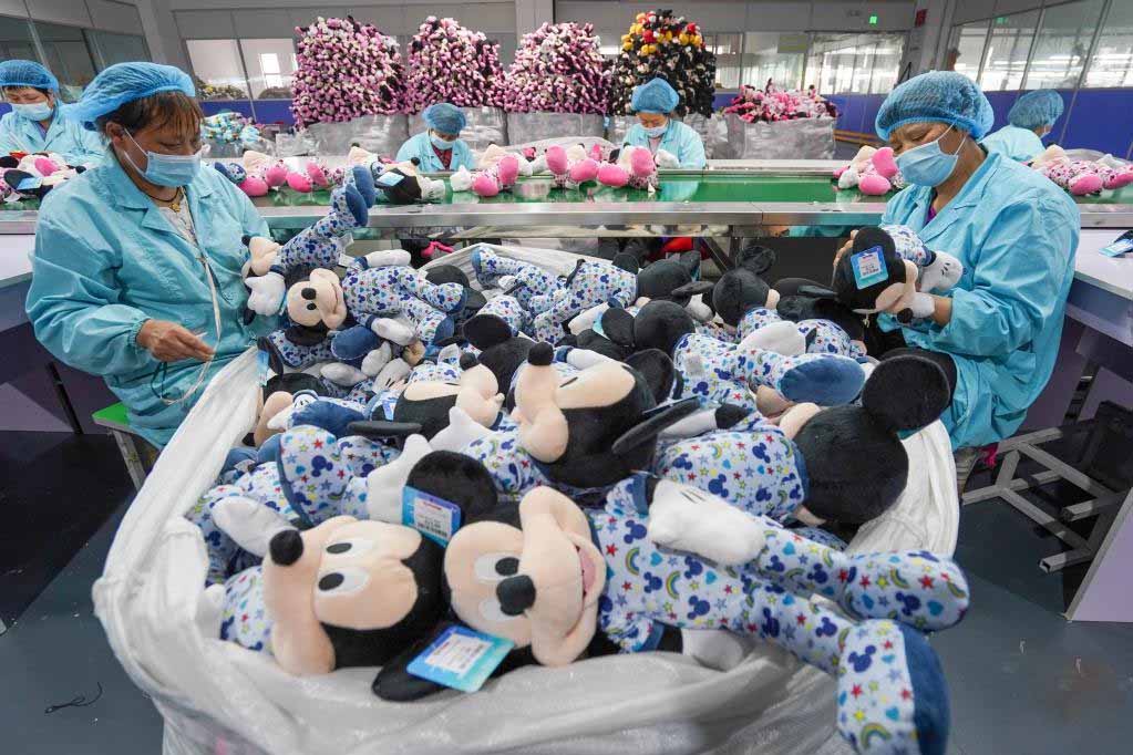 Factory workers in masks and hairnets inspecting Mickey Mouse plush toys during quality control on a production line.