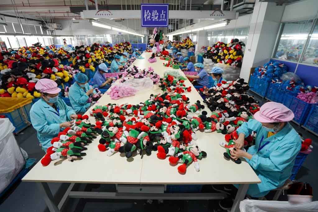 Workers inspecting and sorting stuffed toys at a large factory inspection station with piles of plush animals on tables.