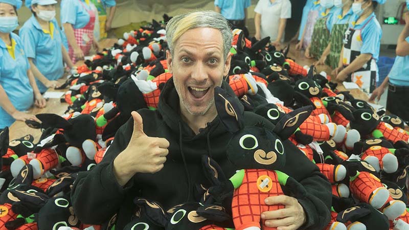 A smiling man gives a thumbs-up while holding a black plush toy in a factory, surrounded by piles of matching plush toys and workers in the background.