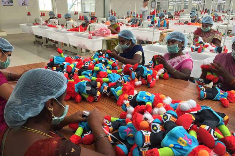 Factory workers wearing hairnets and masks assemble colorful plush toys at long tables in a bright production workshop.