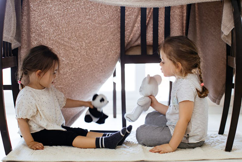 Two young children sitting under a blanket fort, playing with stuffed animal toys on a rug indoors.