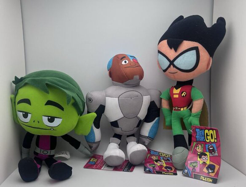 Three Teen Titans plush characters—Beast Boy, Cyborg, and Robin—sitting on a shelf with “Teen Titans Go!” product tags in front.