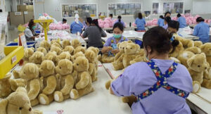 Factory workers inspecting and finishing rows of teddy bear plush toys on tables in a bright production workshop.