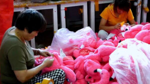 Factory workers sewing and checking pink plush toys in a workshop with bags of stuffed animals.