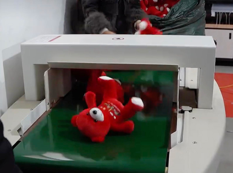 Red plush toy moving through a metal detector tunnel on a conveyor belt during toy factory inspection.