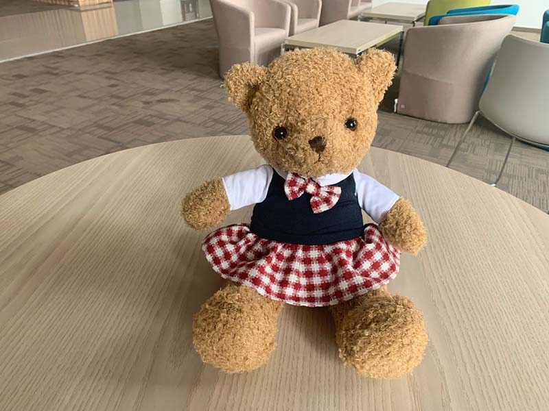 Teddy bear plush wearing a checkered skirt and bow sitting on a round wooden table in a lounge area.