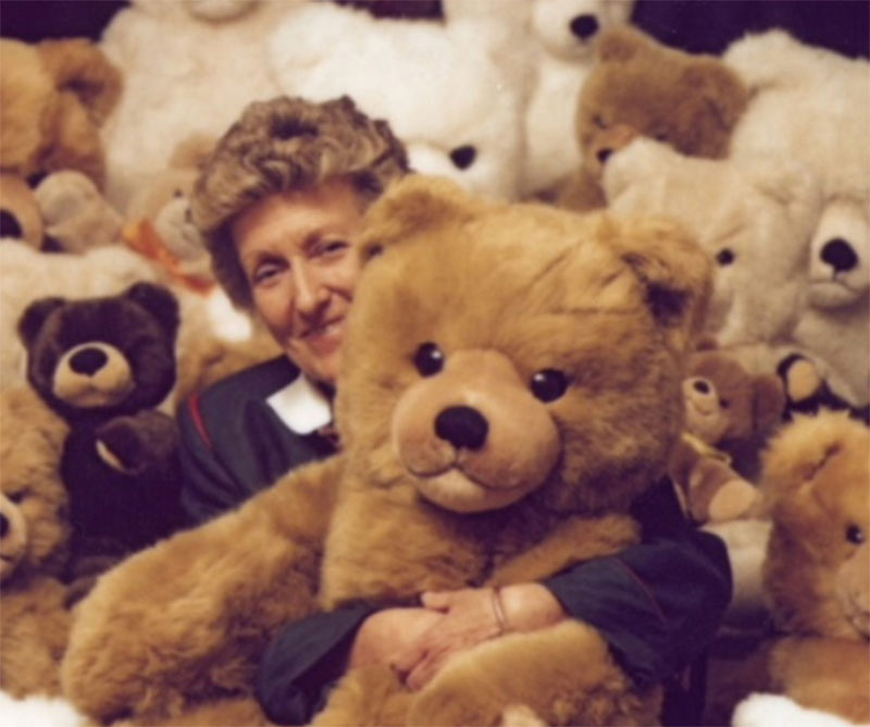 Smiling woman hugging a large teddy bear while surrounded by many plush teddy bears in the background.