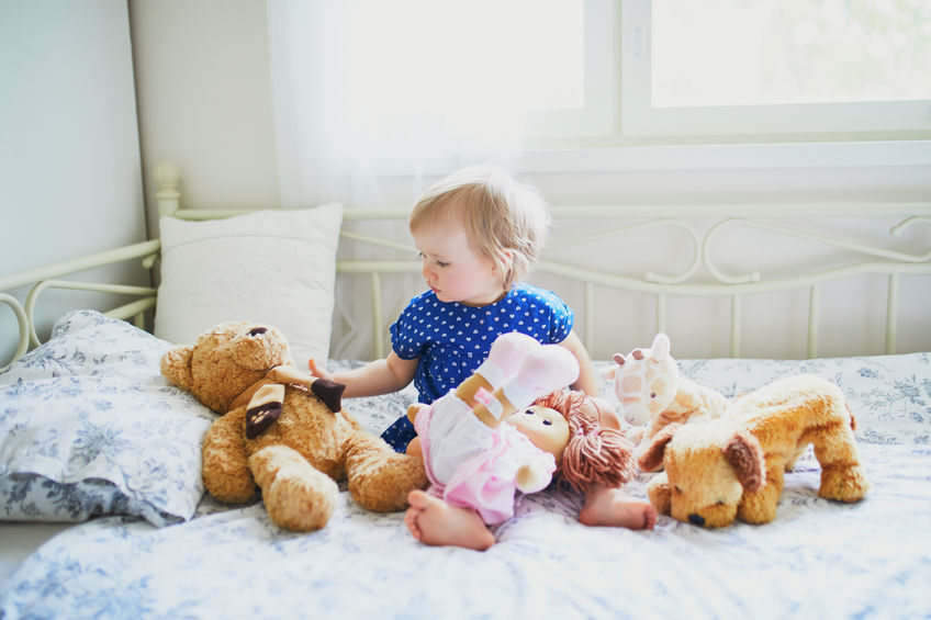 Toddler sitting on a bed, gently playing with stuffed animals in a bright, cozy bedroom