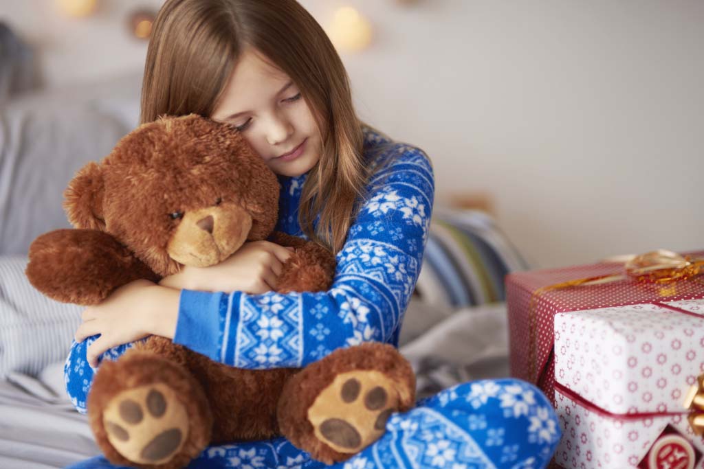 A young child wearing blue patterned pajamas sits on a bed and hugs a brown teddy bear closely, with wrapped gift boxes beside them, conveying comfort, warmth, and emotional attachment to a stuffed toy.
