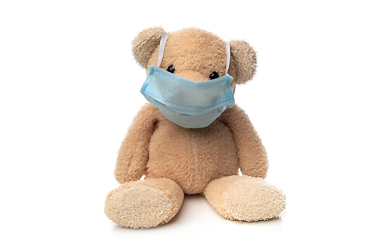 A beige teddy bear sits on a white background wearing a blue surgical face mask, suggesting toy safety and hygiene testing or health-related compliance.