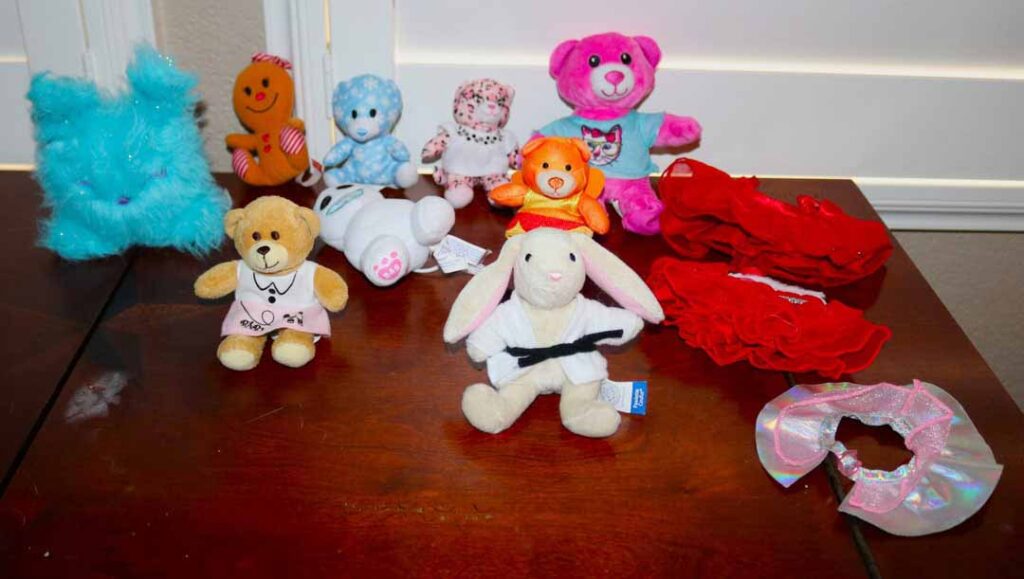 Collection of small Build-A-Bear plush toys and accessories arranged on a wooden table, including teddy bears, a bunny, outfits, and colorful costumes