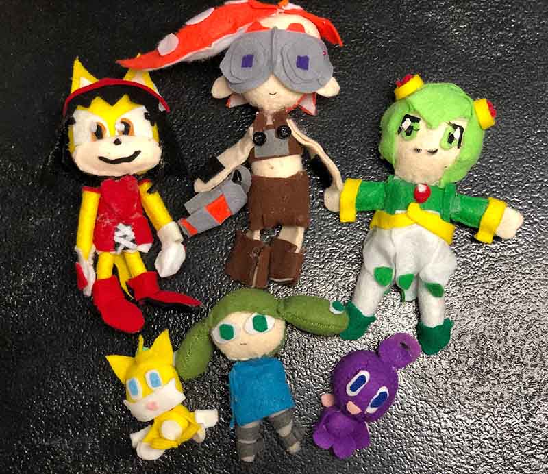 Handmade felt plush dolls arranged on a dark surface, featuring colorful cartoon-style characters with stitched details and simple facial expressions
