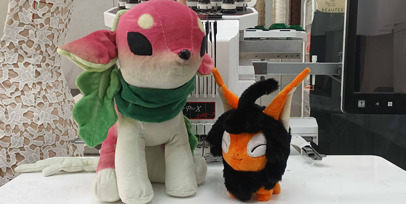 Two custom plush toys displayed on a factory worktable, one pink-and-green deer-like plush with a scarf and leaf details and one small black-and-orange bug-like plush, with embroidery machinery in the background