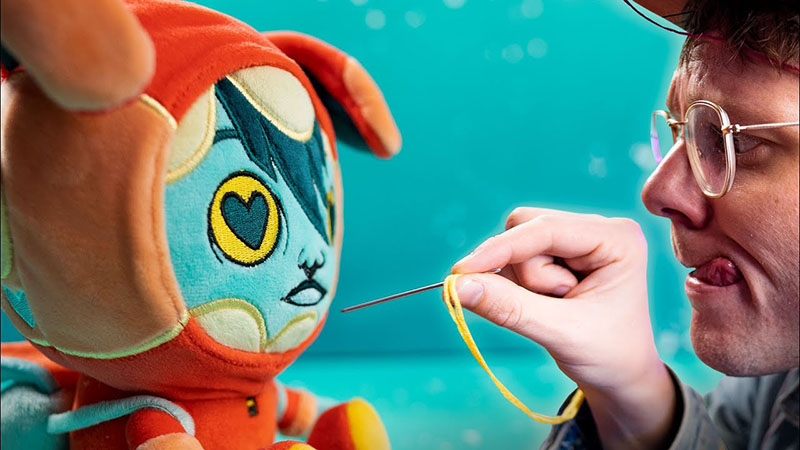Close-up of a plush toy designer hand-stitching facial details on a colorful character plush, carefully guiding a needle and yellow thread while focusing on precise embroidery work