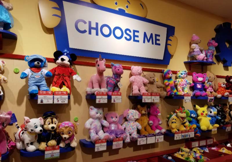 Colorful plush animals displayed on shelves under a “Choose Me” sign at a Build-A-Bear Workshop