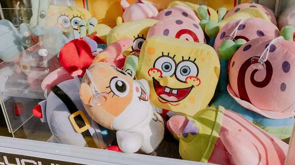 Colorful claw machine filled with cartoon plush toys, including square yellow characters and pink snail-shaped stuffed toys packed closely together