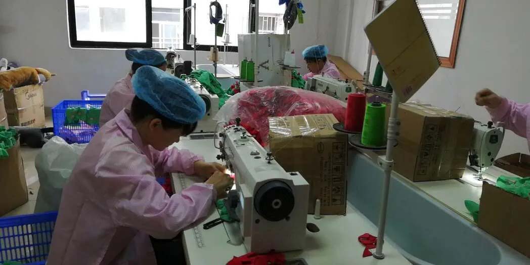 Factory workers operating sewing machines to assemble plush toy components in a production workshop setting.