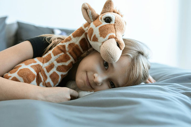 A young child lying on a bed while cuddling a soft giraffe plush toy, highlighting comfort, companionship, and the emotional value of stuffed animals.