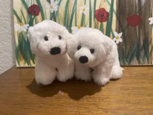 Two soft polar bear plush toys displayed on a wooden surface, featuring fluffy white fabric and simple embroidered facial details.