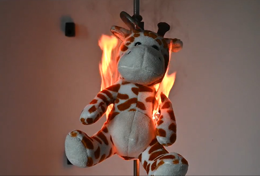 A plush giraffe toy undergoing a controlled flammability test in a laboratory environment, used to evaluate toy safety and compliance with fire safety standards.